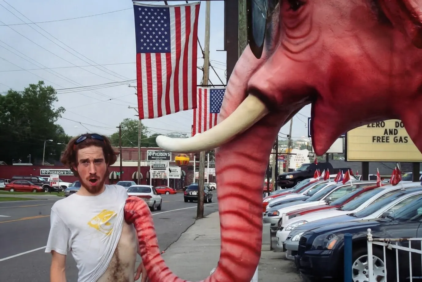 Darby Jones on a sidewalk, looking alarmed, with the trunk of a giant pink fibreglass elephant up his shirt outside a used car lot